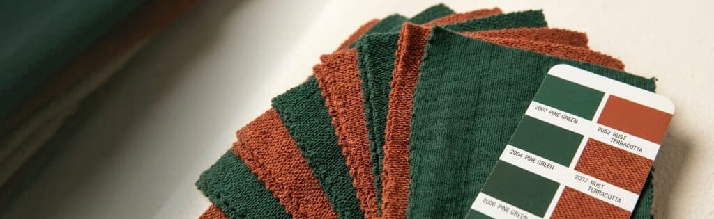 Fanned stack of dyed cotton French terry fabric swatches in matching pine green and rust terracotta, with a Pantone-style color reference card showing the exact dye lot codes
